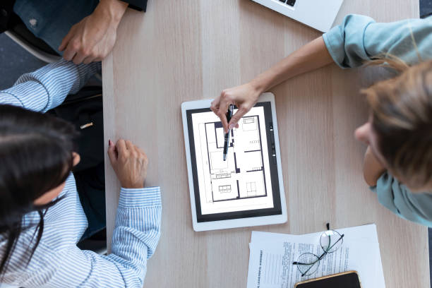 istock Beautiful real-estate agent showing house plans on electronic tablet while talking to the couple about buying the house in the office. 1286567300