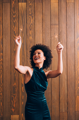 Beautiful smiling woman with an Afro haircut dancing with ignited sparklers in her hands (brown background)