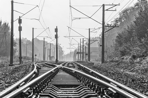 Two railway tracks with electric power poles and cables near a small township's train station. Black and white photo