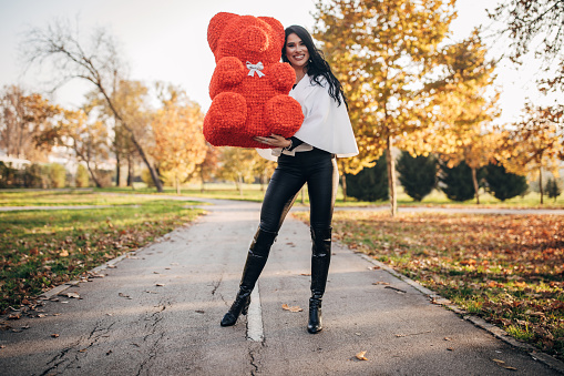 One woman, beautiful young woman holding big red teddy bear in public park.