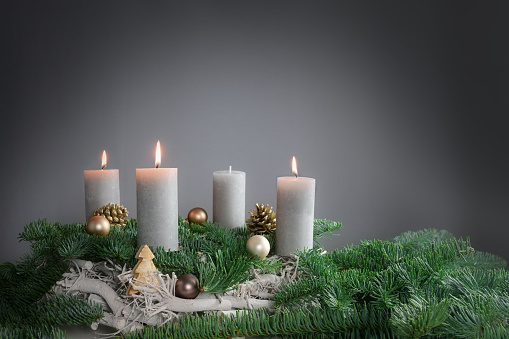 Three of four candles are burning for the third Advent on fir branches with Christmas decoration against a grey background, copy space, selected focus, narrow depth of field