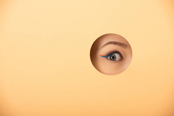 обрезанный вид женщины с подводкой для глаз, глядя на камеру через отверстие на пастельных оранжевый - eye hole стоковые фото и изображения