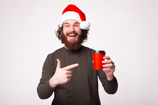 Cheerful young bearded man pointing at red cup of coffee and wearing santa claus hat.