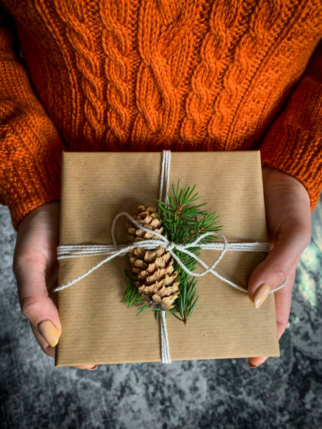 istock Female's hands in pullover holding Christmas gift box decorated with evergreen branch and pine cone. 1286587600