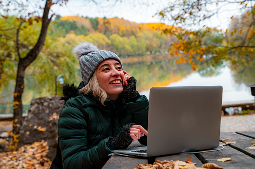 Sonbahar mevsiminde, ormanda gezintiye çıkan kadın göl manzarasında bilgisayar kullanmaktadır. Yaprak döken ağaçlar arasında, sonbahar renklerinden oluşan fotoğraf.