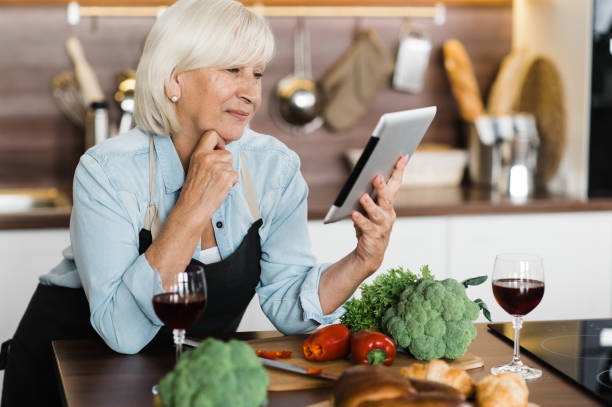 istock Caucasian mature woman in apron standing in kitchen while cooking lunch or dinner and looking at recipe in her tablet 1286592019
