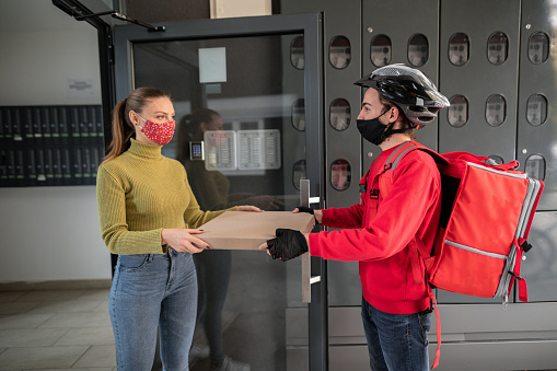Delivery boy delivering pizza to young woman in front of residential building, they are wearing protective face masks for protection against virus during coronavirus pandemic