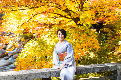 A senior Japanese woman dressed in Kimono looking at camera, enjoying the outdoors during the Autumn season in Japan with beautiful colored Maple leaves from a bridge over a rocky river.