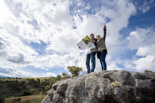 Happy couple of adult Latin American hikers holding a map and pointing at their destination - lifestyle concepts