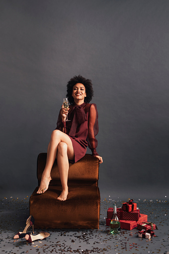 Studio shot of a happy elegant woman with an Afro haircut holding a drink while sitting with her legs crossed; celebration concept