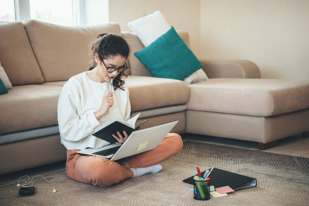 istock Caucasian woman is concentrated on doing homework while sitting on the floor with a laptop and a book 1286615634
