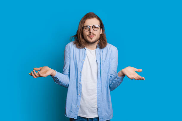 istock Confused caucasian man with beard and long hair is posing indecisive while gesturing with palms on a blue studio wall 1286616360