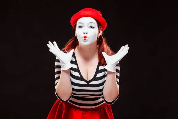 Portrait of female mime artist performing, isolated on black background. Woman in striped clothes and red skirt is standing with raised hands amazed. Symbol of wonder, admiration, worship.