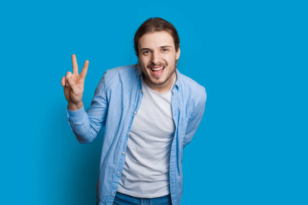 istock Bearded caucasian man is gesturing the hello and peace sign smiling at camera on a blue wall in casual clothes 1286616551