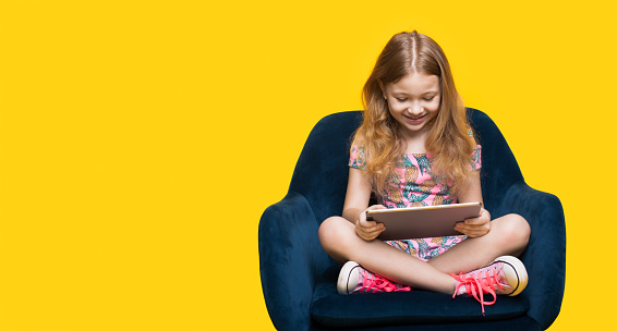 Blonde caucasian girl is playing with a tablet sitting in an armchair on a yellow wall with free space