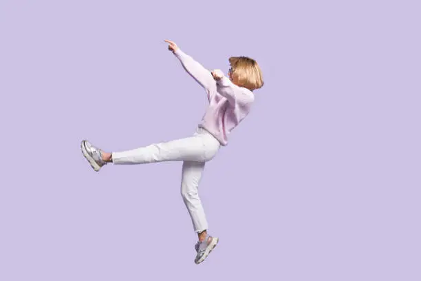 Photo of Caucasian blonde woman is gesturing a falling on a violet studio wall wearing glasses and stylish clothes