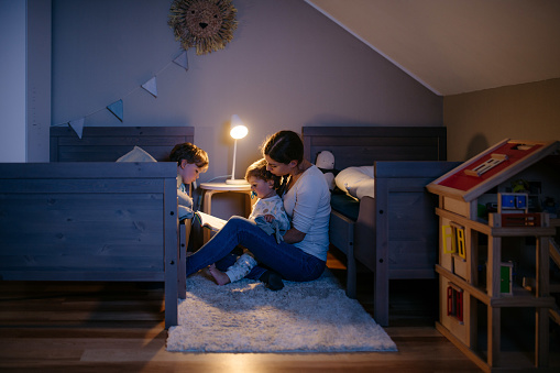 Young mother is reading a fairytale book to her two boys before going to sleep. She is sitting on a carpet next to the beds and reading the book while her two boys are listening. Night table light is switched on. Childrens room with lots of toys. Horizontal photo with copy space.
