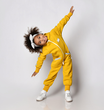 Frolic dark-skinned a curly kid girl in yellow sport jumpsuit performs sideways bands holding arms spread wide like wings, warmup, morning gymnastics in sport school, kindergarten