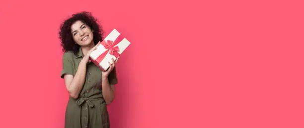 Curly haired woman is posing with a gift smiling at camera on a red wall with free space wearing a dress