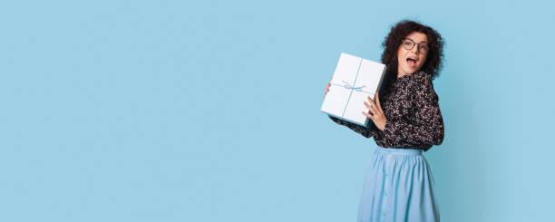 istock Curly haired woman is showing at camera a present box posing happily near blue free space 1286634972