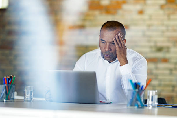 worried black businessman reading an e-mail on laptop in the office. - looking through window businessman problems reading imagens e fotografias de stock