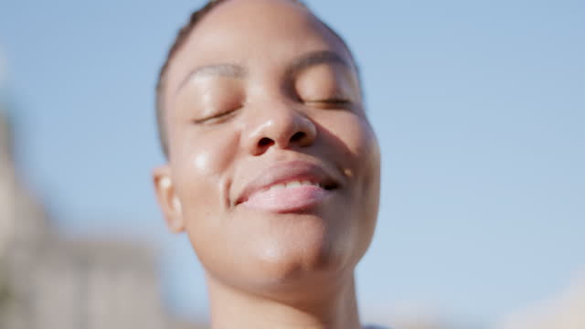 目を閉じた美しい黒人女性のハンドヘルドビデオ映像が、通りでリラックスして晴れた日を楽しんでいます。