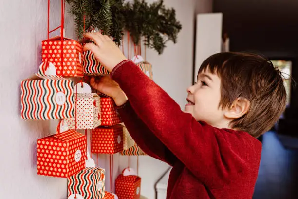 Little happy curious child opening first gift of handmade advent calendar hanging on wall. Sustainable Christmas. Celebrating at home, childhood.