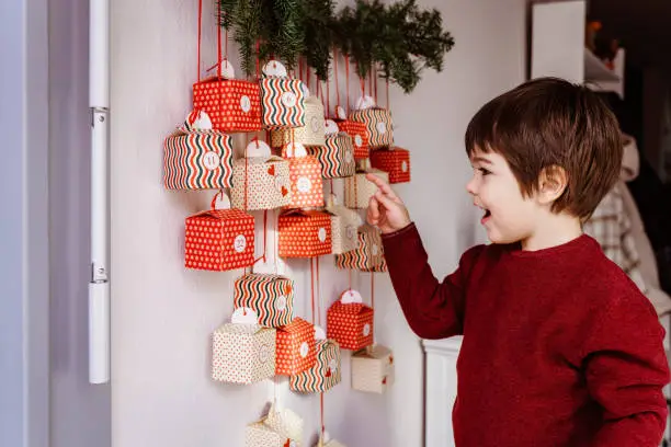 Photo of Happy excited little boy waiting to start opening handmade advent calendar hanging on wall. Sustainable Christmas, upcycling, zero waste, kids seasonal activities