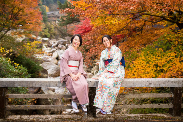 Japanese Mother and Daughter dressed in traditional Kimono and enjoying the Autumn Maple leaves together while sitting on a bridge.