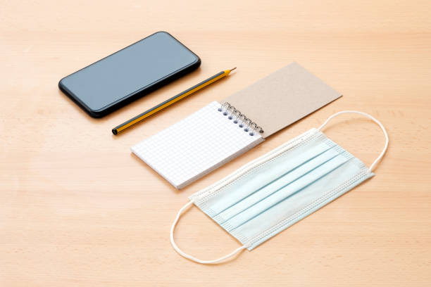 A face mask, a blank spiral notepad with a pen and a smartphone are arranged on a wooden table. To-do list concept during the Covid-19 Outbreak.