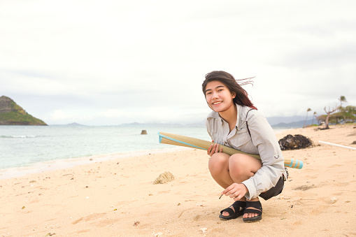 One biracial teen girl sitting on sandy beach in Hawaii playing in the sand with the small island of Mokoli'i or Mokoli’i in background