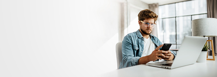 Young man with smartphone in his hands typing message. Modern businessman at sunny office. Freelancer at work. Copy space for text