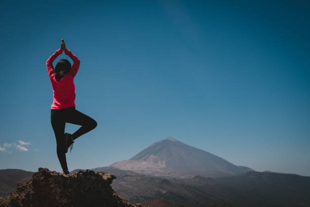 a young woman does yoga on top of a stone looking at a mountain in the background stock photo