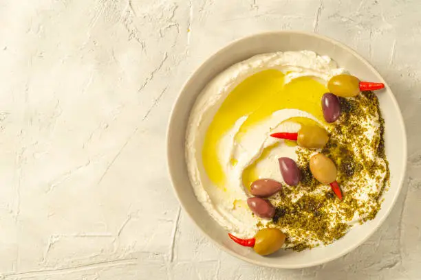 Photo of Popular middle eastern appetizer labneh or labaneh, soft white goat milk cheese with olive oil, hyssop or zaatar, olives on grey table, top view ,flat lay. Close up image.