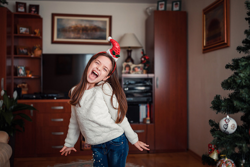 Happy young girl wearing Santa's hat when having fun at home, making funny faces and dancing all over the house