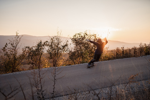 The brave young man chose the extreme way of riding a skateboard down a winding mountain road wearing a protective helmet and gloves on a beautiful sunset