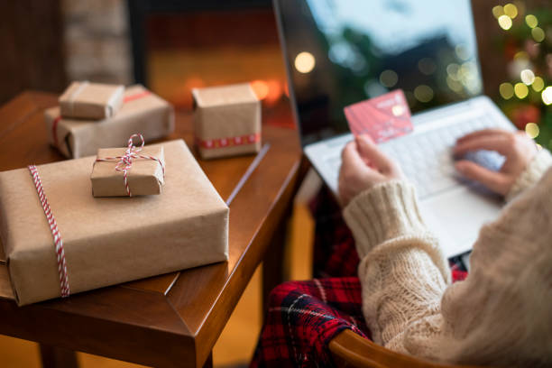 クリスマス。セーターの女性は、暖炉やクリスマスツリーの近くに包装の贈り物でテーブルに座って注文をするためにラップトップを使用してクレジットカードを保持しています。オンライ� - family room ストックフォトと画像