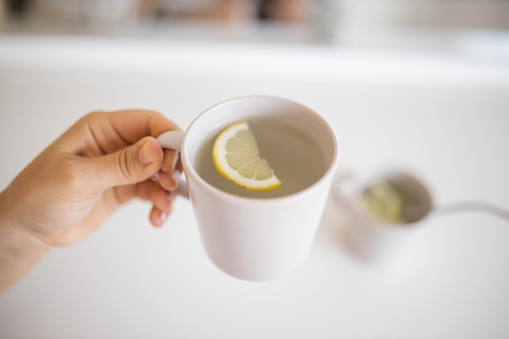 Hand holding cup of lemon tea with lemon slice inside and above blurry cup with a straw. Lemon-flavored drinks in cups on white table. Sweet citric beverages