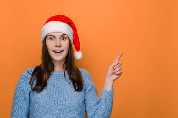 istock Lovely cute woman in red hat points fore finger right, suggests going this direction, demonstrates awesome product, wears sweater, isolated on orange background, has positive smile. Merry Christmas 1286683829
