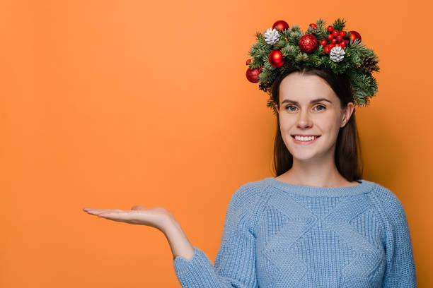 istock Cheerful woman with charming smile keeps palm raised against orange copy space, looks aside, wears Christmas wreath and cozy sweater, demonstrates something. Happy New Year celebration merry holiday 1286684032