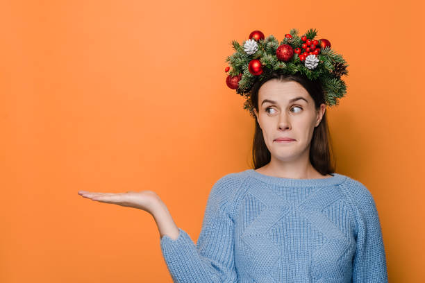 istock Young woman has surprised clueless expression, looks at raised palm in surprise, faces difficult situation, wears handmade wreath and cozy sweater, isolated on orange background. New Year concept 1286684035