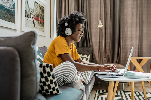 A young African American woman with wireless headphones is using a laptop and working in the living room while smiling and discussing the work on a video call