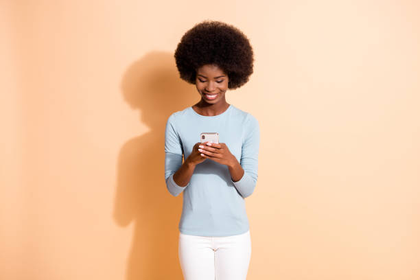 istock Photo portrait of dark skinned curly girl using smartphone browsing internet texting sms in social media isolated on beige color background 1286697049