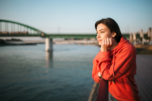 Serene looking young Caucasian woman leaning on the handrails next to a river, looking away in the distance, smiling and day dreaming