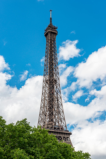 This picture shows the Eiffel Tower from a low-angle viewpoint. The tower, a wrought iron lattice structure, dominates the frame with its wide base narrowing towards the top, where the antenna peaks sharply against the sky. The skies are overcast, with gray clouds suggesting a cloudy day. The foreground has green leafy branches on either side, framing the tower and adding a natural touch to the architectural elegance. It's a perspective that gives a sense of the tower's grandeur and height from a visitor's ground-level viewpoint.