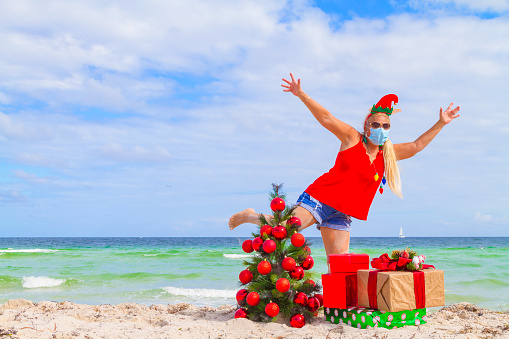 Mature latin beautiful blond fit woman traveling, enjoying the beach, having fun, wearing a Christmas Santa hat, a Christmas lights necklace, a styles sunglasses, showing us her tan and wearing a face cover mask; jumping near to her Christmas tree decorated with Christmas red ornaments at the beach, waiting for opening her presents at the Christmas Day Celebration; on back, the dreaming blue turquoise beach at Miami Beach, Florida, USA during COVID-19 Corona Virus Pandemic Illness Breakdown.