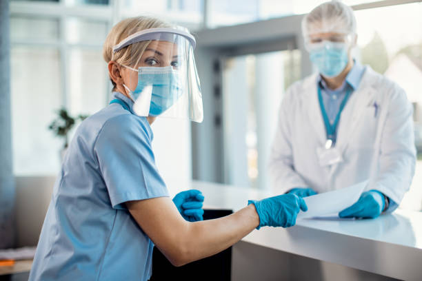 istock Nurse with face shield and mask working at reception desk at the hospital. 1286710559