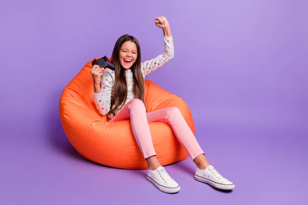 Full length body size photo of little girl playing video games gesturing like winner keeping joystick isolated on bright violet background Full length body size photo of little girl playing video games gesturing like winner keeping joystick isolated on bright violet background. girl sitting stock pictures, royalty-free photos & images