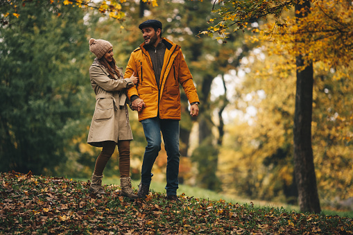 Wife holds her husband by hand and talks to them satisfied that they finally got weekend off. They are walking trough forest, feeling great about Autumn day they are spending.