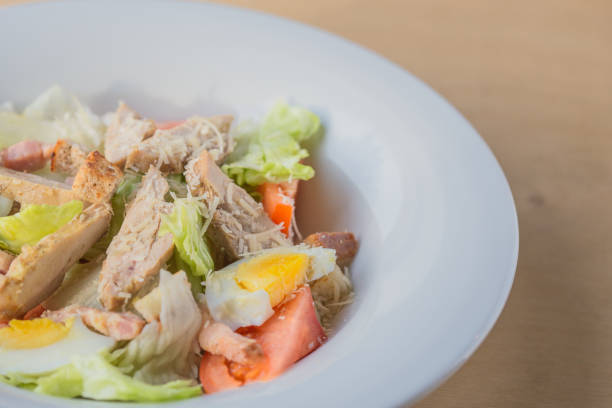 istock Classical Caesar salad with roasted chiken, quail eggs, lettuce leaves, toasts, parmesan, tomatoes and cream sause on the white round plate close up on served table. Italian cuisine 1286712883